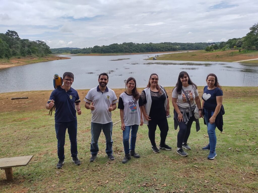 foto de um grupo de 6 pessoas posando com a represa ao fundo. O primeiro homem da foto tem uma arara no ombro.